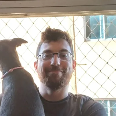Douglas, de óculos e barba, com um cachorro no colo em frente a uma janela.